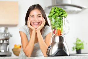 Woman in kitchen with vegetables in blender smiling, illustrating that Grand Avenue Smiles can restore your beautiful smile.