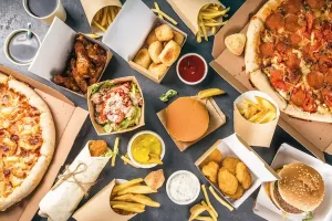 A top-down, flat-lay view of an assortment of fast food spread across a dark gray surface. The spread includes two large pizzas in cardboard boxes, multiple burgers, fried chicken wings, chicken nuggets, a wrap, various containers of French fries, a small salad, and several dipping sauces like ketchup and mustard.
