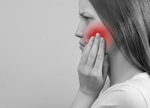 Black and white photo of a young woman from the side, touching her cheek with a red glow, illustrating the concept that Grand Avenue Smiles provides emergency appointments.