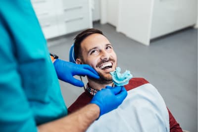 A man with a beard sits in a dental chair, smiling and looking up at a dentist. The dentist, wearing teal scrubs and blue gloves, holds a blue dental impression mold near the patient's mouth. The setting is a clean, modern dental office.