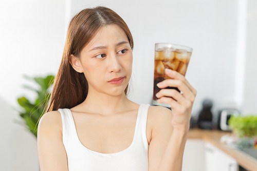 Young Asian woman looking at a glass of cola, illustrating the concept that diet drinks are also bad for your teeth.